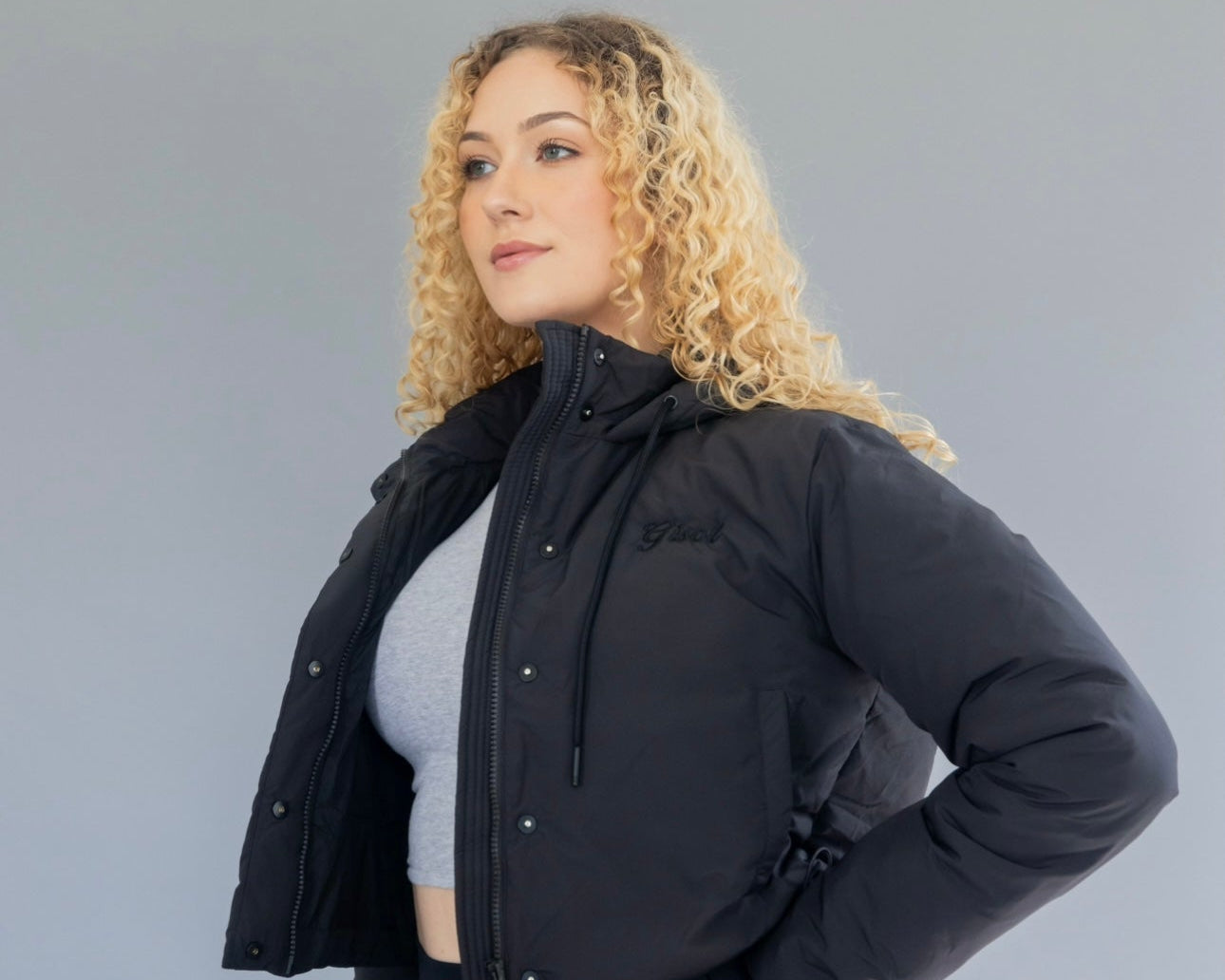 A woman modeling a waist-length black puffer jacket with a hood, featuring button and zipper front closure, adjustable drawcord at the hood and waist, and side pockets.